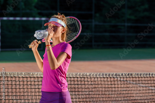Close up cute young girl in sporty cap drinking water from plastic bottle after hard tennis training on the outdoor court background at sunset.