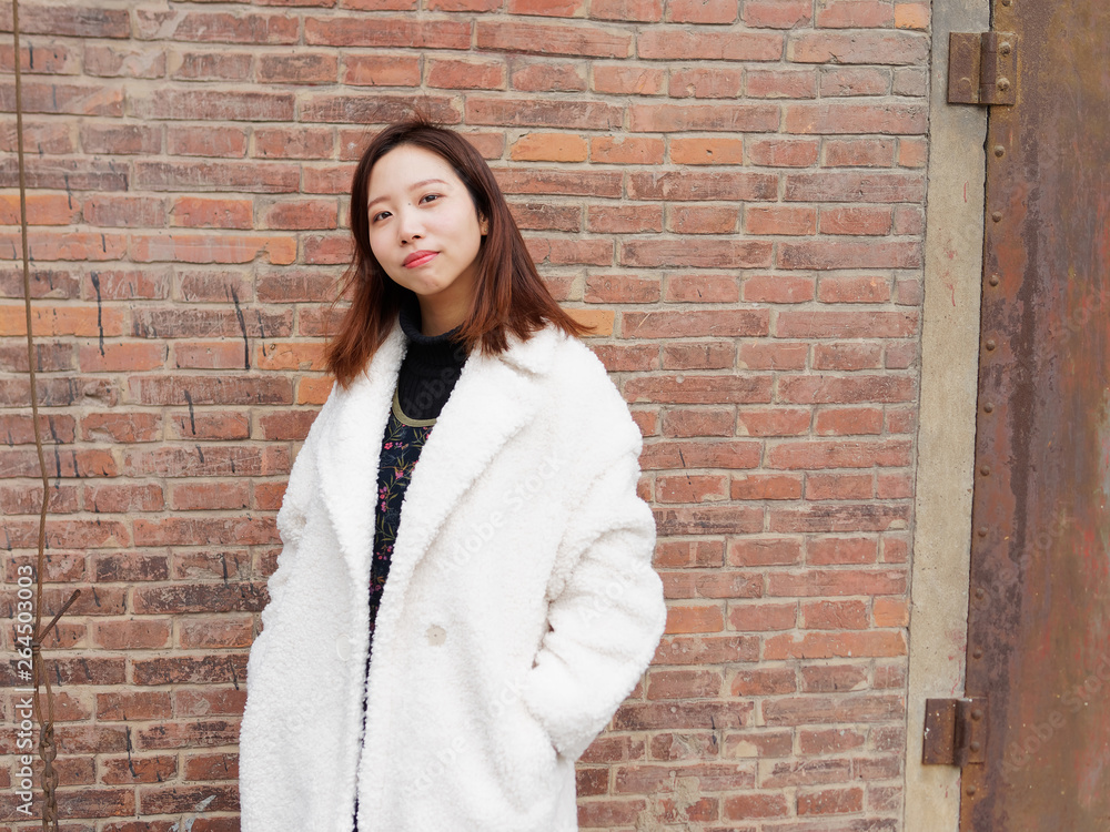 Street photography of a beautiful Chinese woman in white coat with red brick wall background, looking at you with two hands in her pocket, portrait of cute girl with nevus near eyes. Street photography of a beautiful Chinese woman in white coat with red brick wall background, looking at you with two hands in her pocket, portrait of cute girl with nevus near eyes.