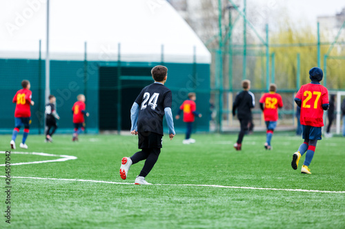 Boys at black red sportswear run, dribble, attack on football field. Young Soccer players with ball on green grass. Training, football, active lifestyle for kids 