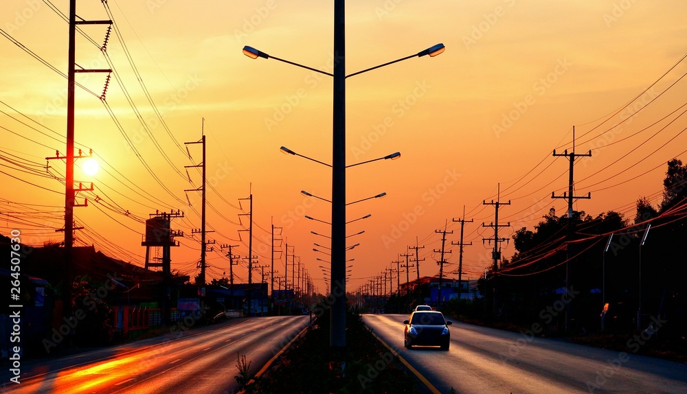 Silhouette row of street light posts and electric poles with power lines and car driving on the road against colorful sunrise sky background in perspective view