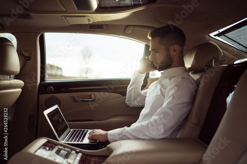 Man uses laptop and talking on smartphone sitting in back seat of car, businessman in taxi © D'Action Images