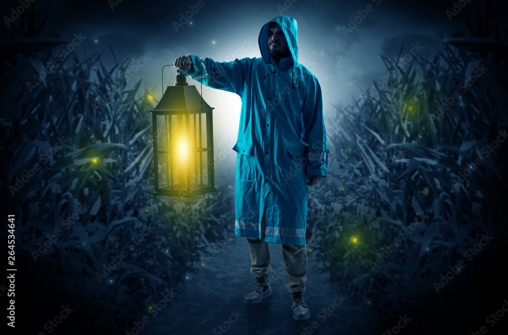Fototapeta premium Man in raincoat at night coming from thicket and looking something with glowing lantern