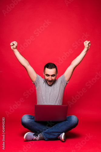Portrait of a joyful young man looking at laptop computer while sitting on a floor and celebrating isolated over red background