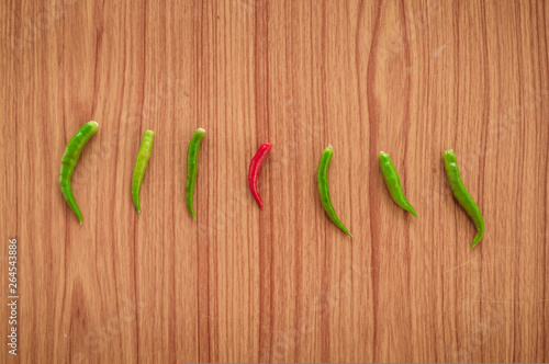 A Red hot spicy thai chili peppers or Mirchi among green chilli arranged in a horizontal row on wooden background, flat lay. Top view. Standing Out From The Crowd Concept with copy space room for text