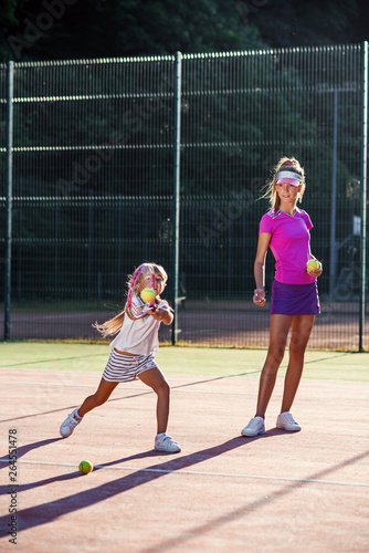 Little girl in white uniform hitting ball under net during tennis training with coach. Attractive female coach making exercises with little tennis player on the outdoor court at sunset.