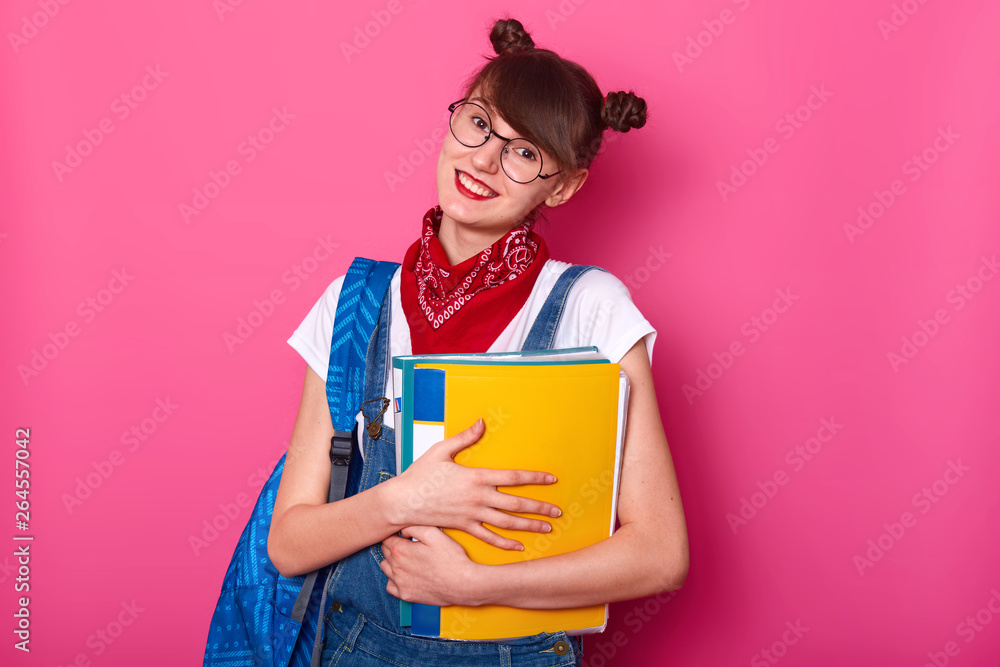 Happy schoolgirl with paper folder isolated on rosy background. Smiling girl being glad to come back school after summer holidy. Lady wears t shirt and overalls, tilts her head tio side and smiles Happy schoolgirl with paper folder isolated on rosy background. Smiling girl being glad to come back school after summer holidy. Lady wears t shirt and overalls, tilts her head tio side and smiles