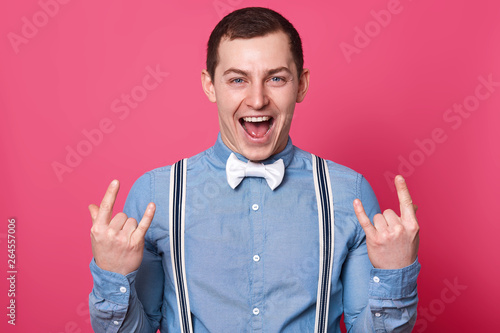 Portrait of joyful happy man showing rock gesture against pink studio background, standing with enjoying facial expression, screaming something, looks cool, wears shirt, bowtie and suspenders.