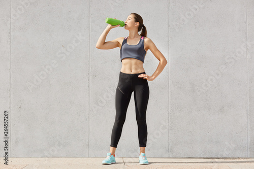 Athletic woman in sportswear drinking water from plastic container while having workout in gymnasiun, keeps hand on hip. Full body length portrait isolated on gray studio background. Healthy lifestyle