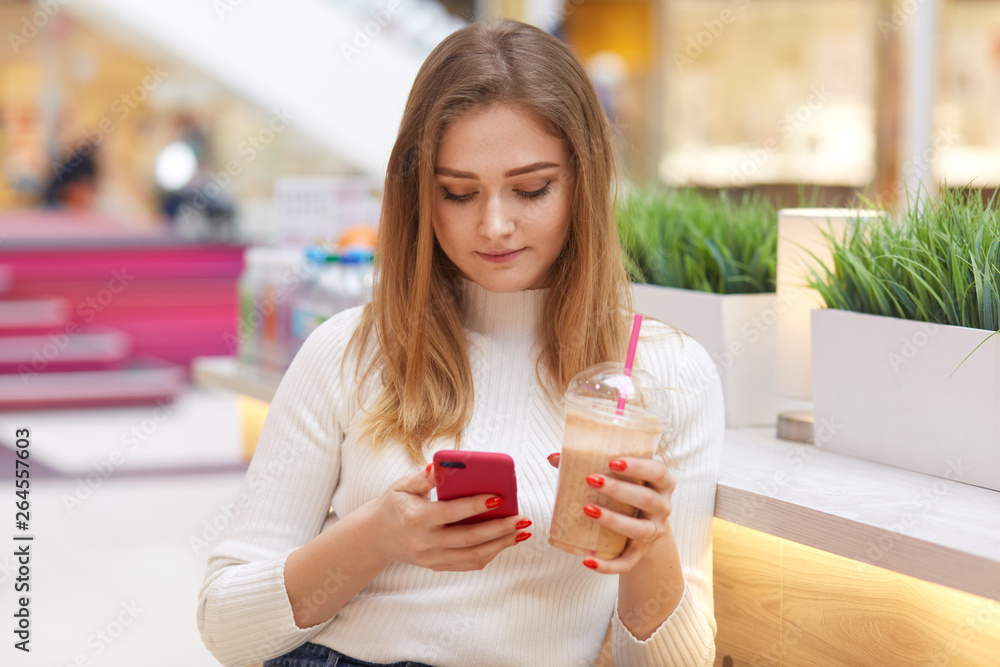 Beautiful blonde student sitting in cafe, reading text message on her smart phone and drinking lemonade from takeaway plastic cup, dressed casually, comunicates with friend, uses wireless Internet. Beautiful blonde student sitting in cafe, reading text message on her smart phone and drinking lemonade from takeaway plastic cup, dressed casually, comunicates with friend, uses wireless Internet.