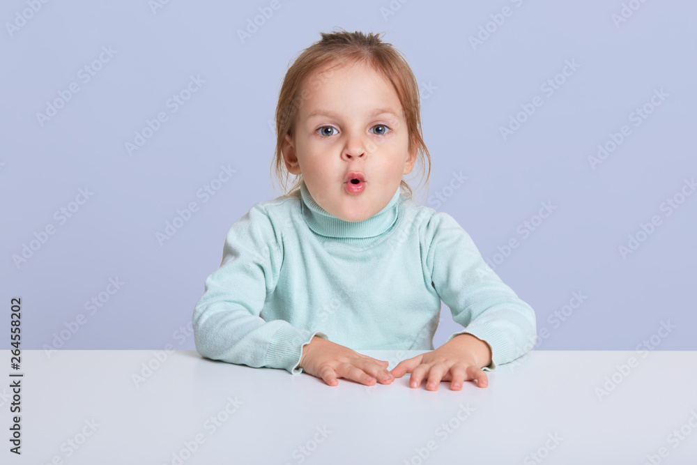 Image of astonished lovely little sitting at white desk, small female child with rounded lips looking with surprised expression, posing isolated over blue background with copyspace for advertisment. Image of astonished lovely little sitting at white desk, small female child with rounded lips looking with surprised expression, posing isolated over blue background with copyspace for advertisment.