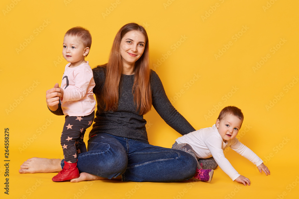 Studio shot of young mother and her twin toddlers poses in photo studio isoleted over yellow background. Mommy sits with her infants on floor and hugs them with great love. Motherhood concept. Studio shot of young mother and her twin toddlers poses in photo studio isoleted over yellow background. Mommy sits with her infants on floor and hugs them with great love. Motherhood concept.