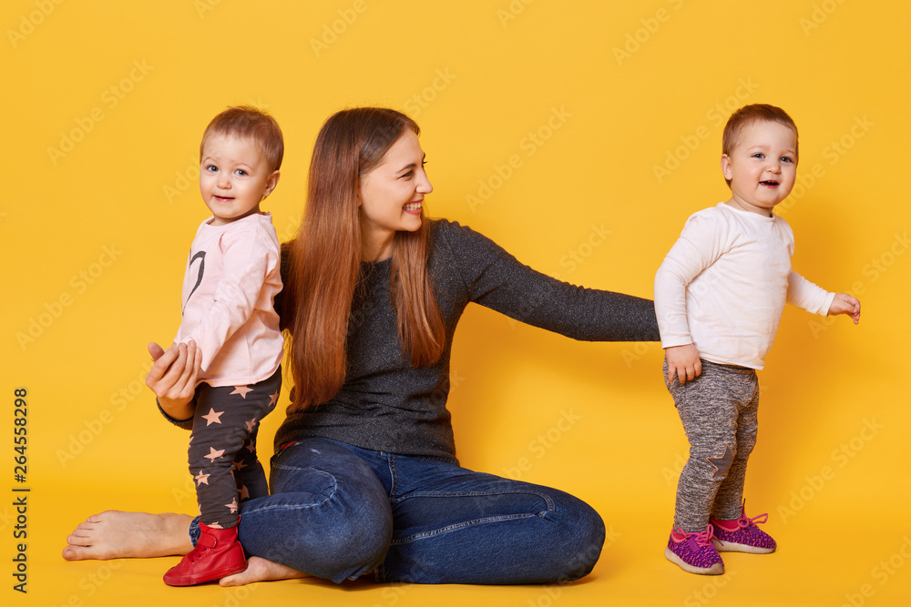 Image of attractive brown haired mother wants to be photographed with her sweet children, sit on floor in studio. Mother and girls twins, dressed casually, mom spends time with her doughters. Image of attractive brown haired mother wants to be photographed with her sweet children, sit on floor in studio. Mother and girls twins, dressed casually, mom spends time with her doughters.