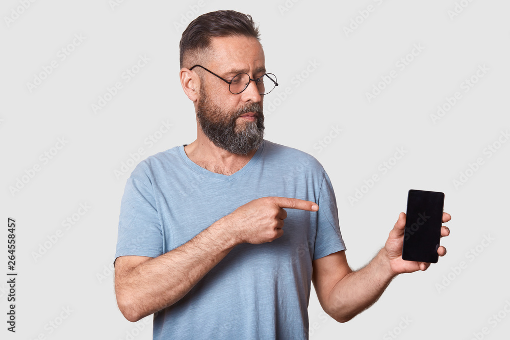 Close up portrait of attractive middle aged male in gray casual t shirt, stands against white wall, pointing with fore finger on blank screen of his smartphone, looking on device. Copy space. Close up portrait of attractive middle aged male in gray casual t shirt, stands against white wall, pointing with fore finger on blank screen of his smartphone, looking on device. Copy space.