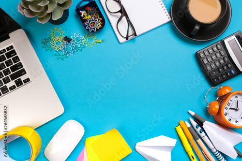 Office desk table with set of colorful supplies, white blank note pad, cup, pen, pc, crumpled paper, flower on blue background. Top view and copy space for text