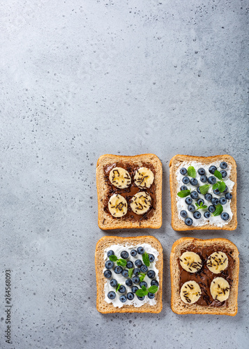 Delicious open sandwiches with goat cheese and blueberries and chocolate and banana on gray table. Breakfast background. Copy space, top view