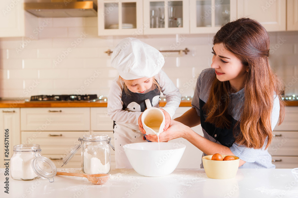 The little daughter in the chef's hat and apron and her mother prepare baking in the bright, classic kitchen.