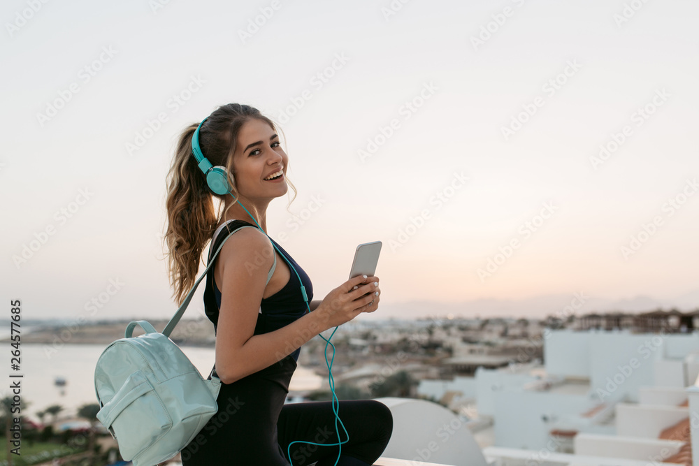 Enjoying lovely music through headphones of positive joyful sportswoman chilling on seafront on sunrise background. Fashionable model, having fun., smiling to camera. Place for text