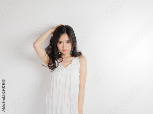 Portrait of beautiful Chinese woman in sexy white dress posing with white wall background, young girl puts her hand on head and looks unhappy.
