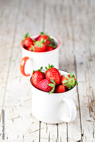 Organic red strawberries in two white ceramic cups on rustic wooden background.