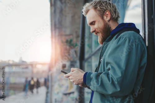 Busy young man stand outside on street. He hold phone in hands and smile. Guy lean to wall. Sunset and shining. Evening.