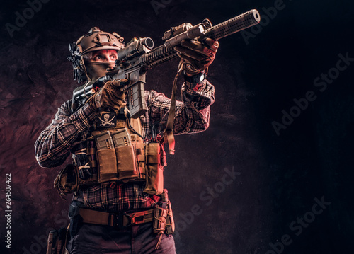 Elite unit, special forces soldier in camouflage uniform holding an assault rifle with a laser sight and aims at the target. Studio photo against a dark textured wall