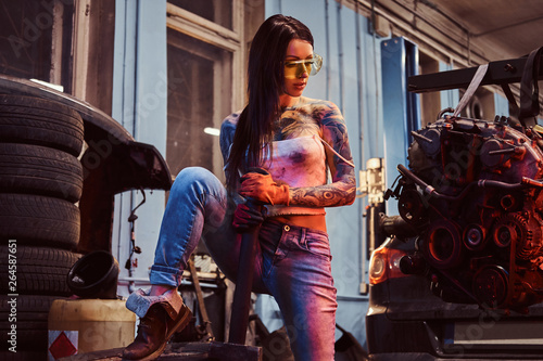 Female model with tattooed body wearing protective goggles posing with a steel hammer next to a car engine suspended on a hydraulic hoist in the workshop. Photo with red light illumination