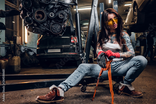 Stylish tattooed girl holding a big wrench while sitting on a hydraulic hoist with a suspended car engine in the workshop
