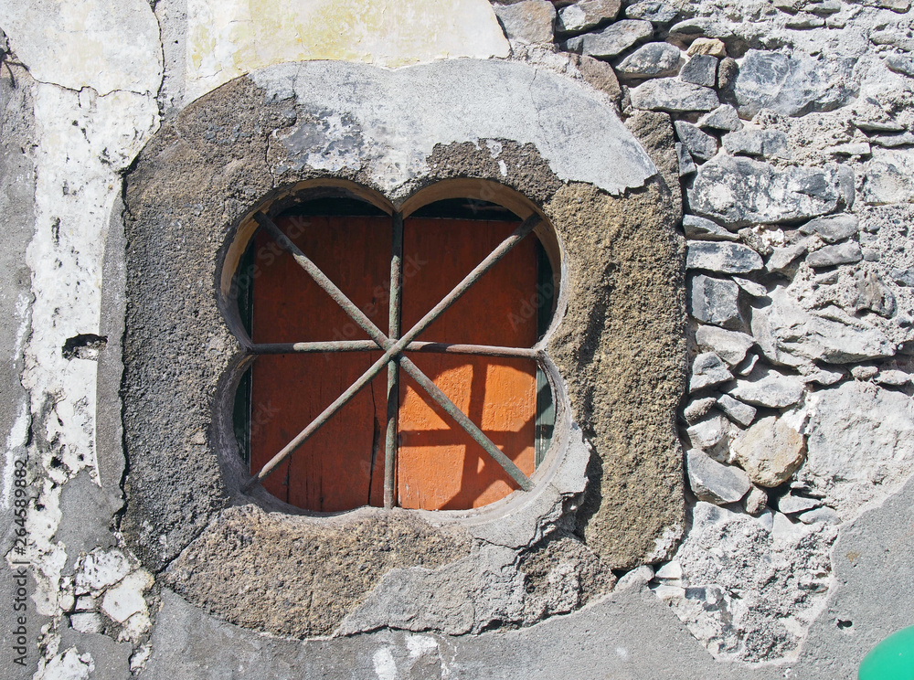 and ancient stone oblong shaped window with crossed bars with an orange shutter set in an old patched repaired grey wall with cracked cement