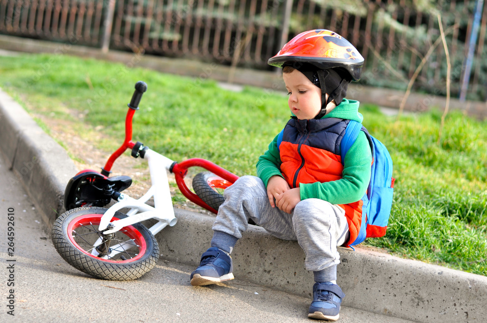 Fototapeta premium Sports Kid boy in a protective helmet sits on the curb near his red white balance bike