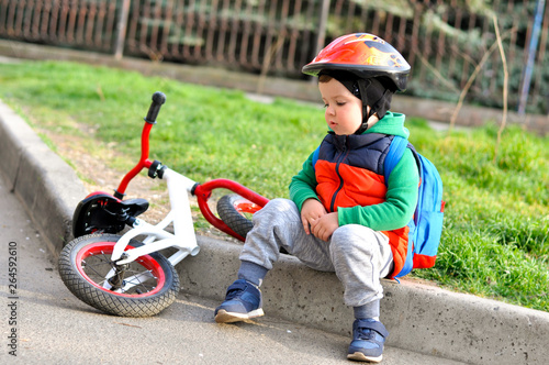 Sports Kid boy in a protective helmet sits on the curb near his red white balance bike