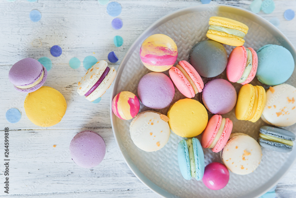 Multicolored french macaron cakes on blue plate and white wooden background. White, yellow, pink, purple and grey french macarons with fresh berries. Multicolored french macaron cakes on blue plate and white wooden background. White, yellow, pink, purple and grey french macarons with fresh berries.