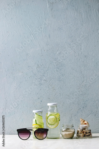 Summer theme. Two glass bottles with lemon and cucumber infusion sassy water, seashells and sand in glass jars, sunglasses on white marble table, concrete wall at background.