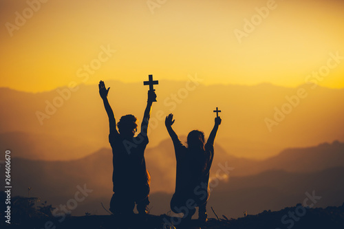 Young male and girl sitting holding christian cross for worshipping God at sunset background. christian silhouette concept.
