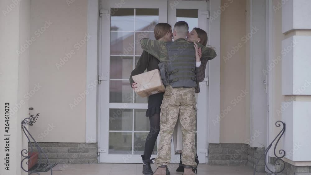 The young soldier in military closes running to his family, cuddling his sisters holding paper with welcome home signature and gift box in front of house. A man returns from the military service The young soldier in military closes running to his family, cuddling his sisters holding paper with welcome home signature and gift box in front of house. A man returns from the military service