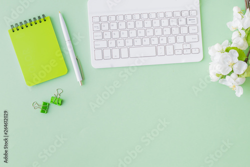 Flat lay blogger or freelancer workspace with a notebook, keyboard and white spring flowers on a green background