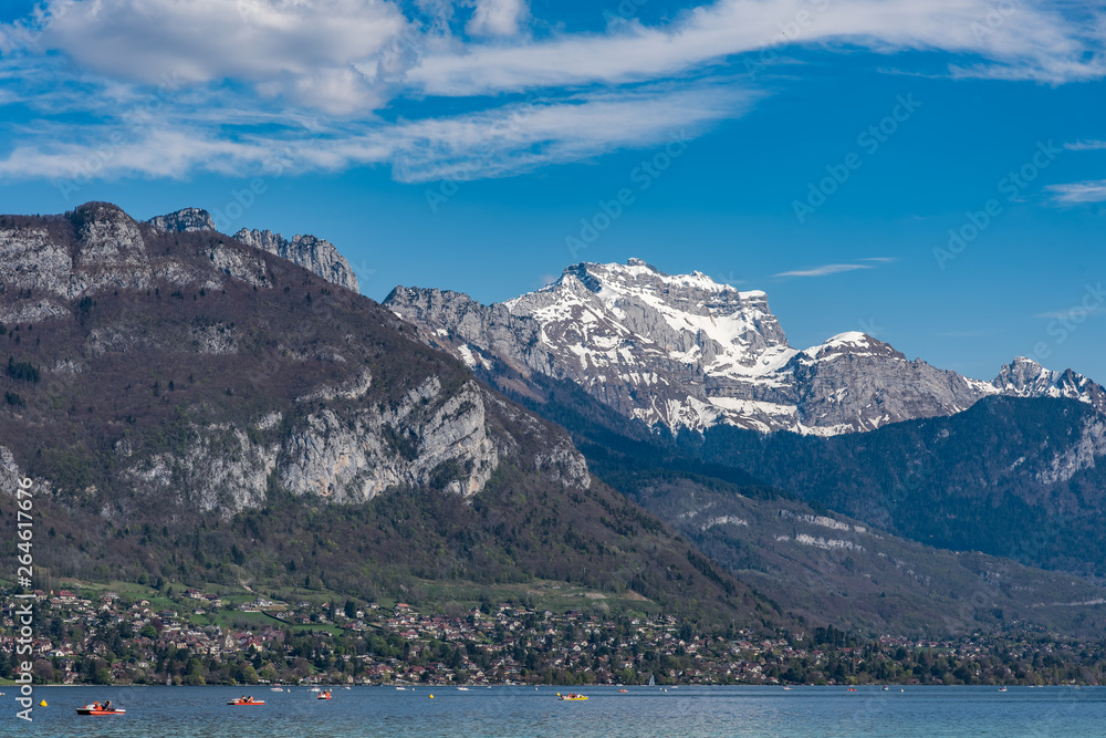 A sunny day on an Alpine lake. There are small pleasure boats on the lake. Beyond the water there is a village and green fields, and a snow-covered mountain peak. It is Lake Annecy, France.