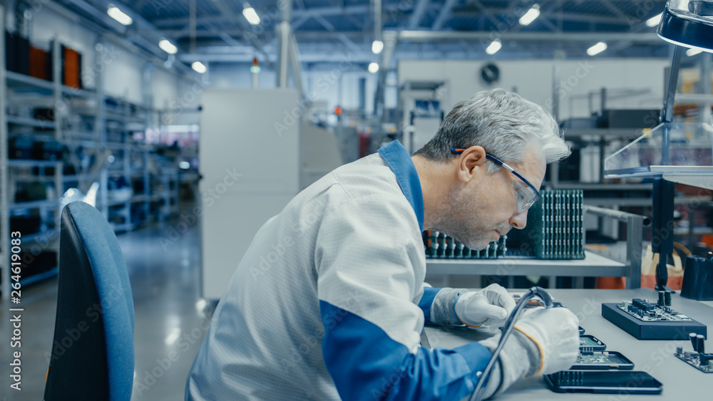 Senior Man in Blue - White Work Coat is Using Plier to Assemble Printed Circuit Board for Smartphone. Electronics Factory Workers in a High Tech Factory Facility.