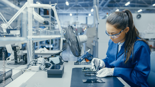 Young Female Blue and White Work Coat is Using Plier to Assemble Printed Circuit Board for Smartphone. Electronics Factory Workers in a High Tech Factory Facility. photo