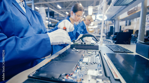 Close-Up of a Female Electronics Factory Worker in Blue Work Coat Assembling Laptop's Motherboard with a Screwdriver. High Tech Factory Facility with Multiple Employees.  photo