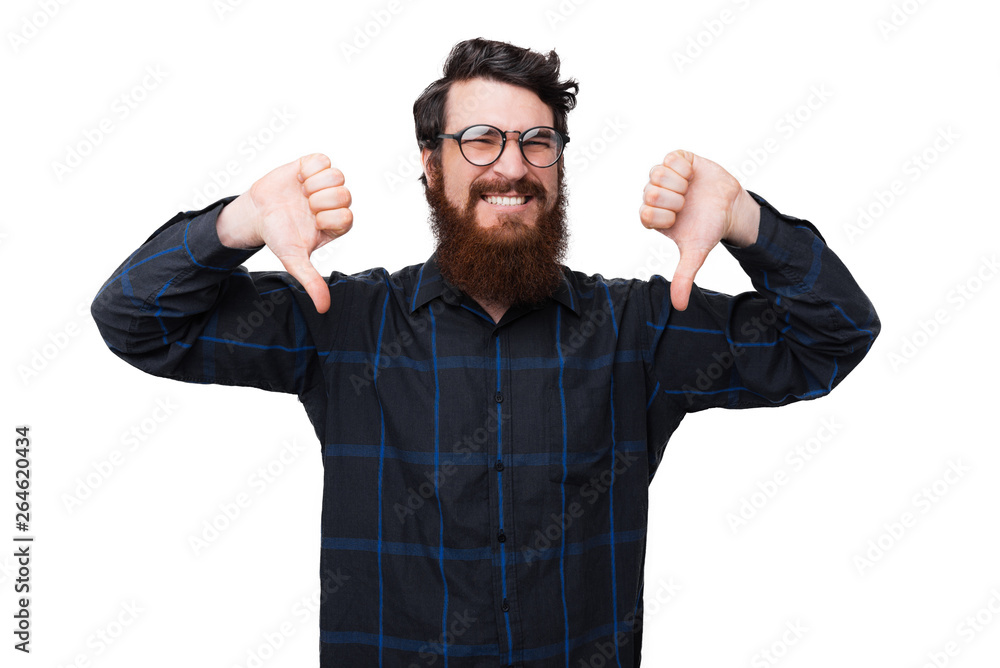 Fototapeta premium A bearded nerd, showing a dislike gesture with thumbs down over white background