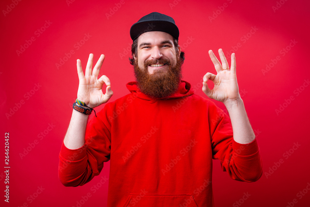 A cheerful bearded guy in red hoody, showing ok sign with both handsand looking at camera A cheerful bearded guy in red hoody, showing ok sign with both handsand looking at camera