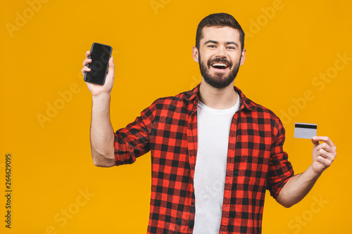 Portrait of a smiling young man holding blank screen mobile phone and showing credit card isolated over yellow background.