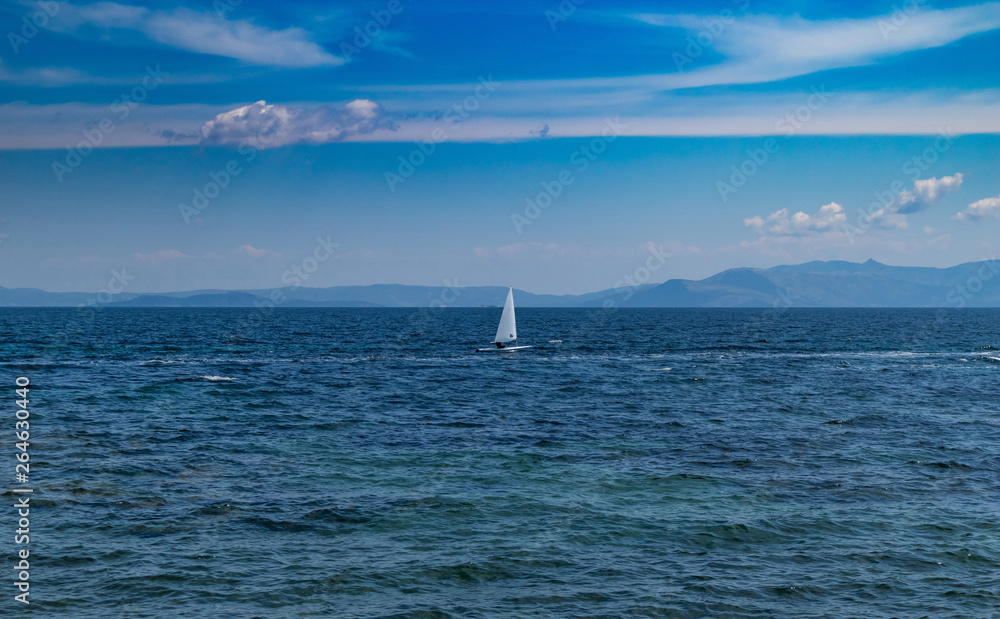 Small optimist boat with white sail, blue sky and sea background