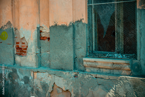 Orange old building with very weathered plaster with a wooden framed broken window with rusty metallic grid over it – Weak construction in ruins with a damaged facade © madrolly