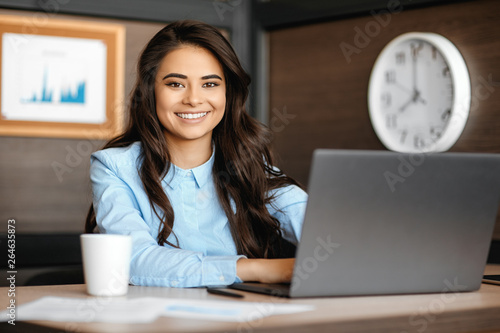 Photo of beautiful young manager woman with bright smile working with laptop in modern office.