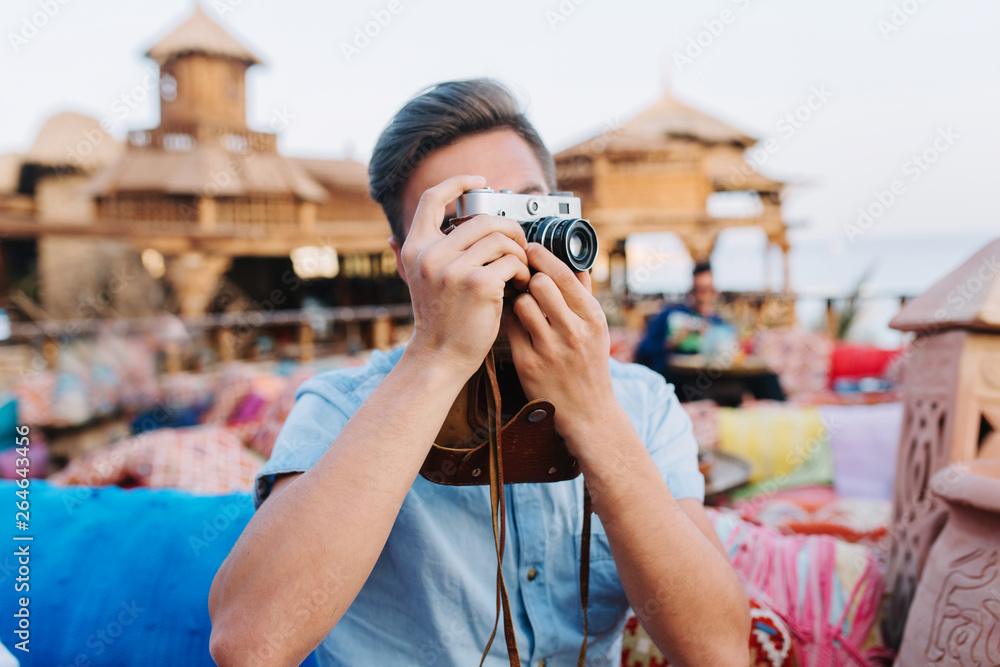 Young man with professional retro camera resting in outdoor cafe, waiting for friends. Guy wearing trendy denim shirt making photo of city view, sitting in open-air restaurant in summer morning