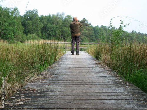 Birdwatcher (man) standing on a boardwalk in Dutch nature reserve. Looking at a bird on the lake. Concept for future, discovery, exploring and education.
