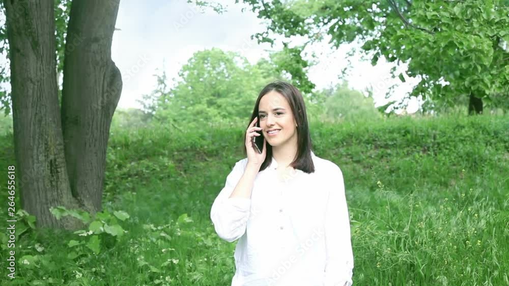 Happy cute caucasian young woman smiling and talking on the phone in the park. Atractive girl in white shirt with cellphone Happy cute caucasian young woman smiling and talking on the phone in the park. Atractive girl in white shirt with cellphone