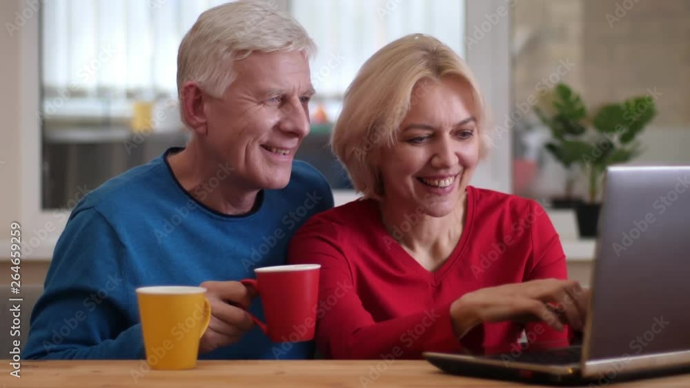 Closeup shoot of senior happy couple networking on the laptop on the desk and drinking tea indoors in a cozy apartment Closeup shoot of senior happy couple networking on the laptop on the desk and drinking tea indoors in a cozy apartment