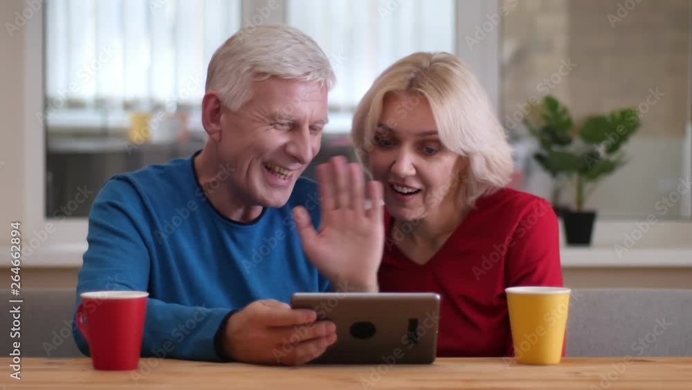 Closeup shoot of aged happy couple having a video call the tablet with cups with tea on the desk indoors in a cozy apartment Closeup shoot of aged happy couple having a video call the tablet with cups with tea on the desk indoors in a cozy apartment